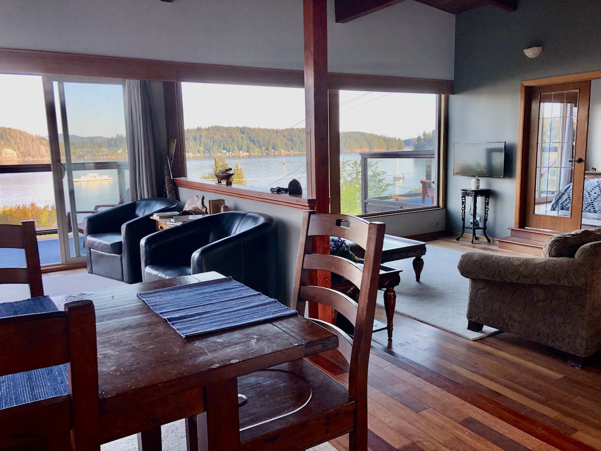 The living area features large windows that offer stunning views of the water and surrounding landscape. Comfortable seating includes two black armchairs and a light-toned sofa. A wooden dining table is positioned nearby, complimented by light blue place settings.