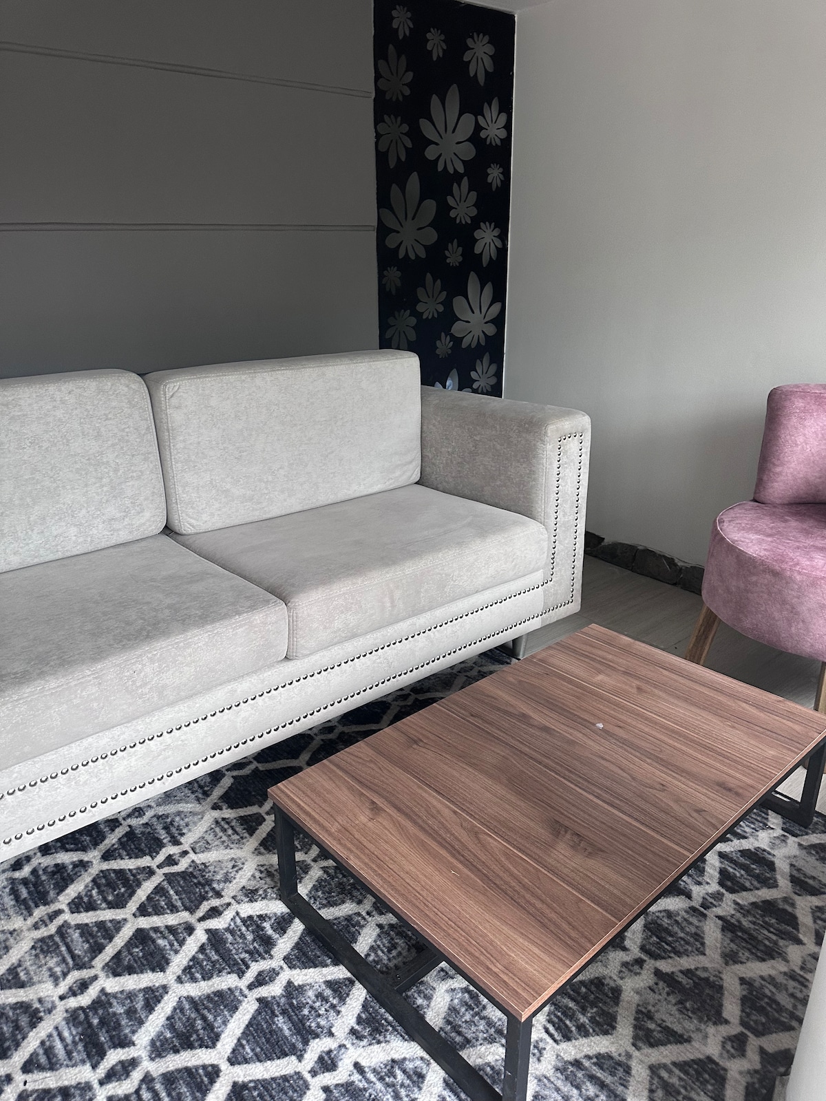 A light gray sofa is positioned beside a wooden coffee table on a patterned rug. A pink accent chair complements the seating area, while a wall adorned with floral wallpaper adds a decorative touch to the space.