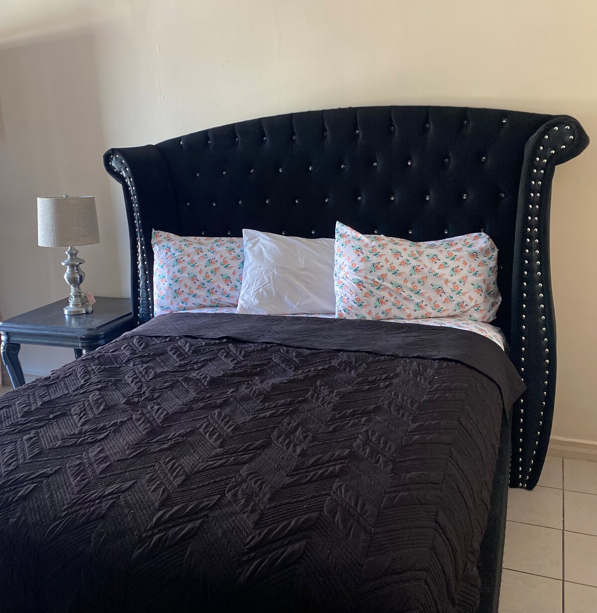 A bed features a tall, tufted black headboard adorned with silver studs. Soft, floral-patterned pillows rest against the headboard, complementing a dark, textured blanket draped over the bed. A small bedside table with a lamp is positioned nearby.