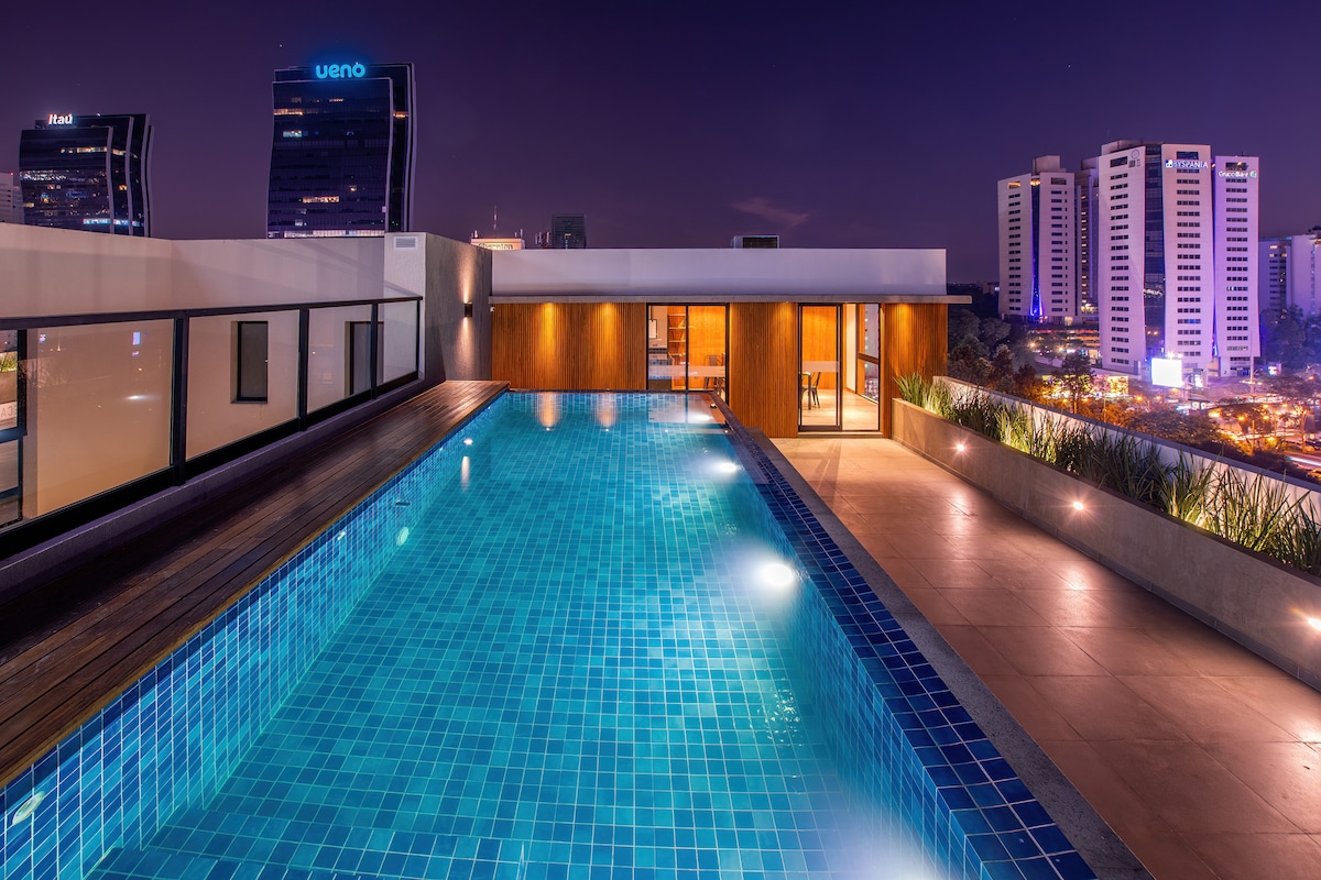An infinity pool reflects the city skyline at night, framed by wooden decking. The serene water surface is illuminated, creating a calming atmosphere. Modern buildings are visible in the background, offering a glimpse of the urban landscape surrounding the rooftop area.