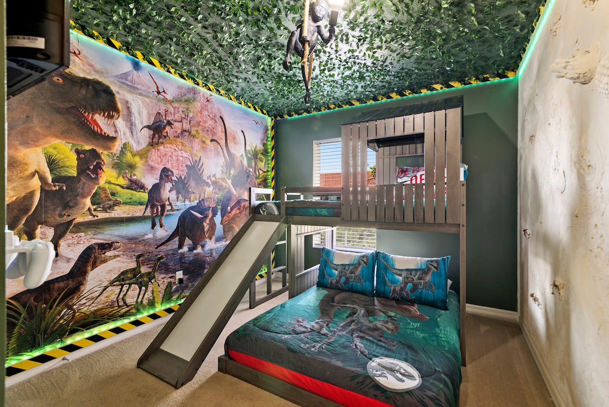 A themed children's bedroom features a loft bed with a slide, decorated with a dinosaur mural across the ceiling and walls. The bedding displays dinosaur graphics, while bright LED lighting enhances the playful atmosphere. A window allows natural light to fill the space.