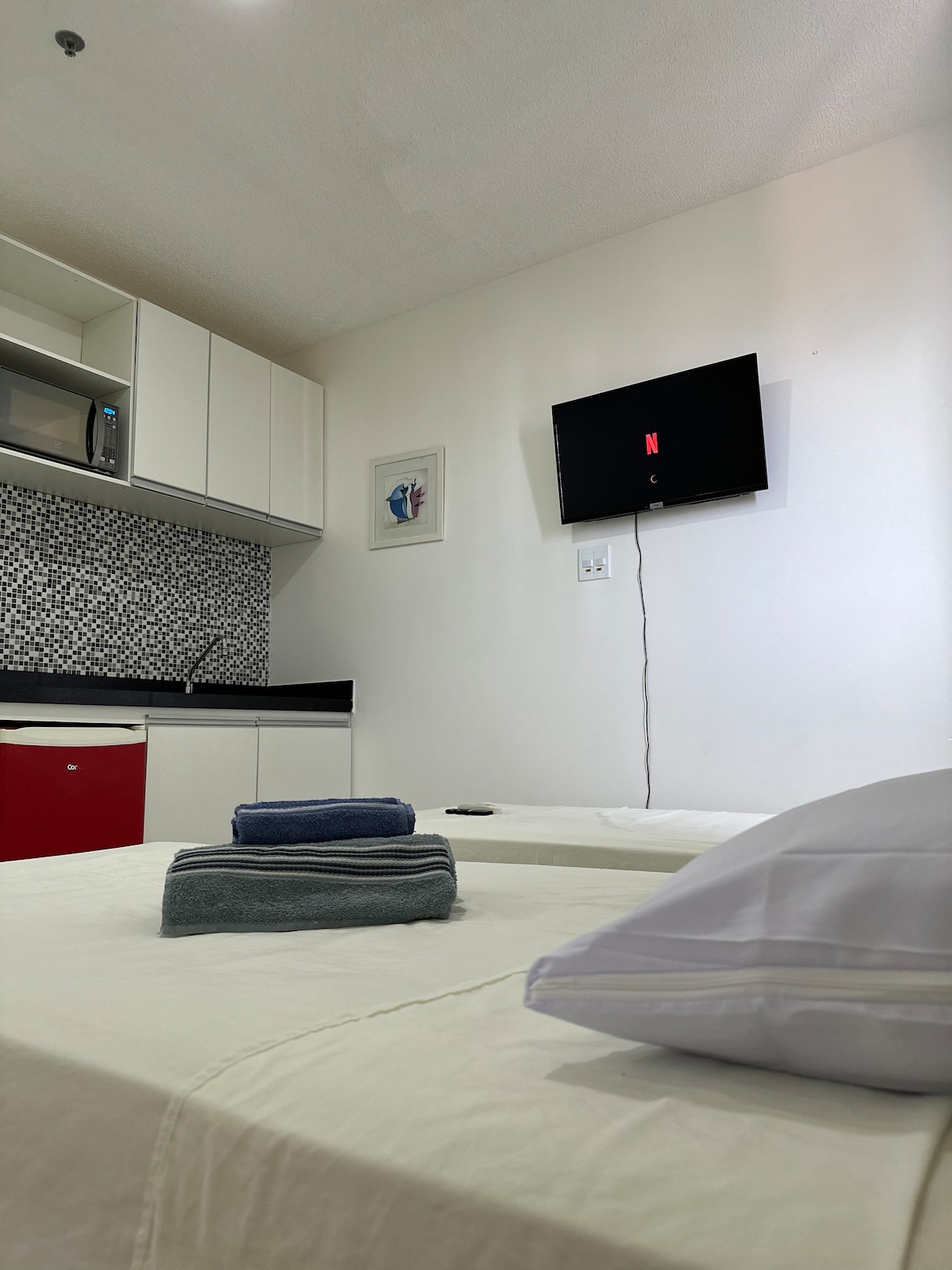 A cozy room features a spacious bed topped with neatly arranged towels. A Smart TV is mounted on the wall, displaying the Netflix logo. In the background, a compact kitchen area with a microwave is visible, along with a patterned backsplash and individual cabinets.