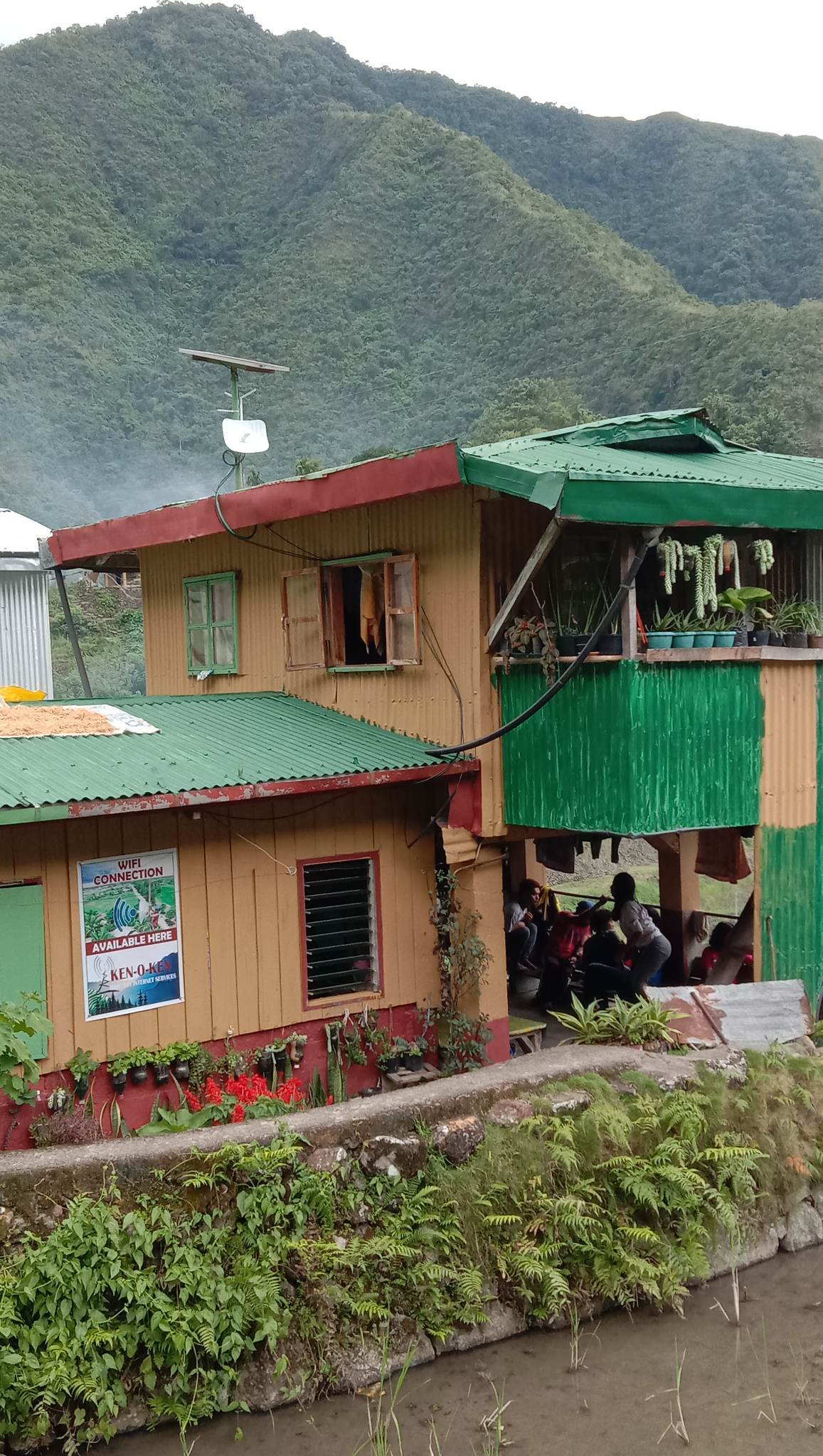 A two-story building constructed with colorful wooden panels is nestled against a mountainous backdrop. Green roofs complement the beige walls, while windows allow a glimpse of the vibrant indoor space. Surrounding greenery enhances the natural setting of the location.