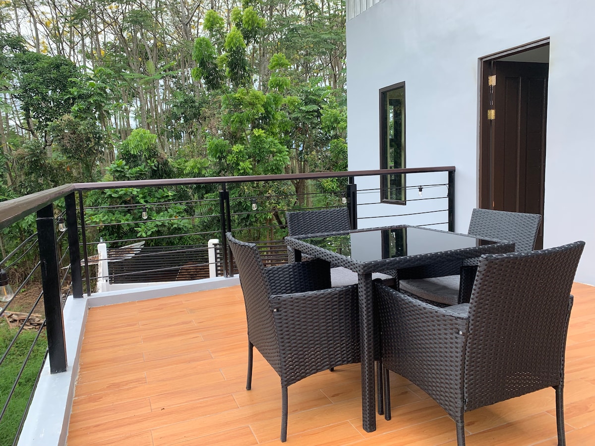 A private balcony is equipped with a glass-top table and four woven chairs, offering a serene space for relaxation. Lush greenery can be seen beyond the railing, providing a natural backdrop that enhances the outdoor setting.