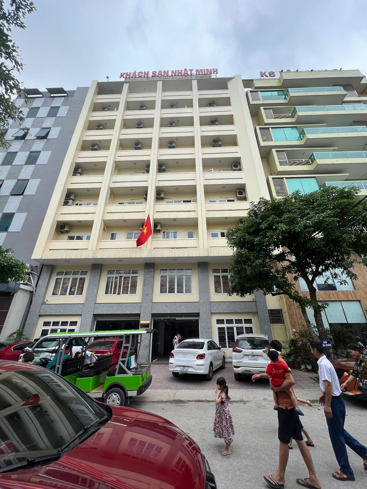 A six-story building stands prominently, displaying the name 'Khách Sạn Nhật Minh' on its rooftop. The facade features large windows and air conditioning units. A public parking space is visible in the foreground, with several parked vehicles and pedestrians nearby.