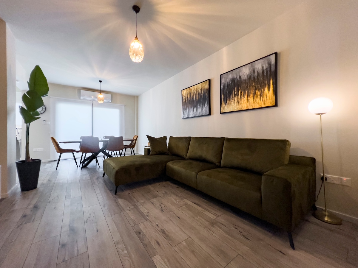 A modern living area is presented with a large olive green sectional sofa facing two abstract art pieces. A floor lamp stands nearby, and a dining table with chairs is positioned in the background, adjacent to a window allowing natural light to illuminate the space.