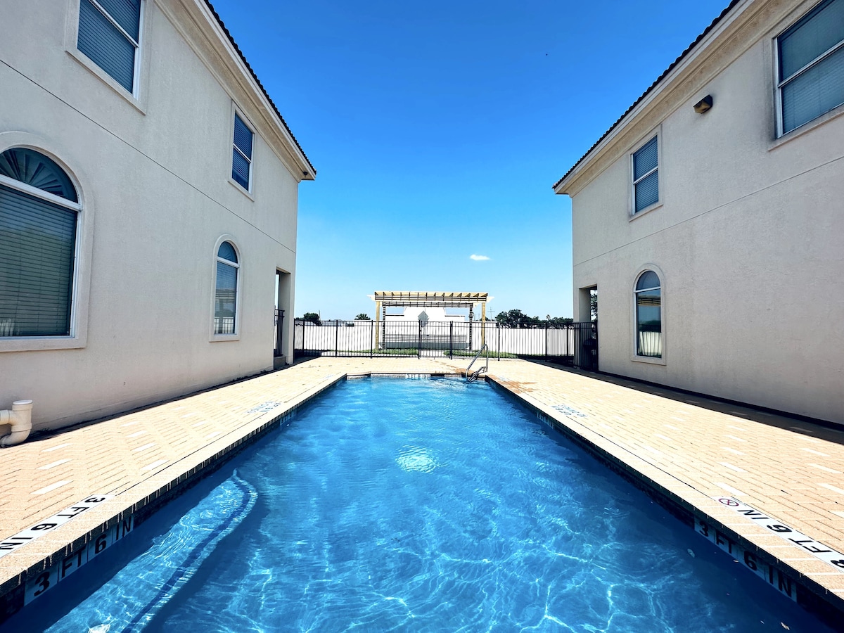 A serene pool area is framed by two light-colored buildings, featuring a clear blue water surface. A trellis structure is positioned at the end of the pool, surrounded by a spacious sun deck, inviting relaxation under the open sky.