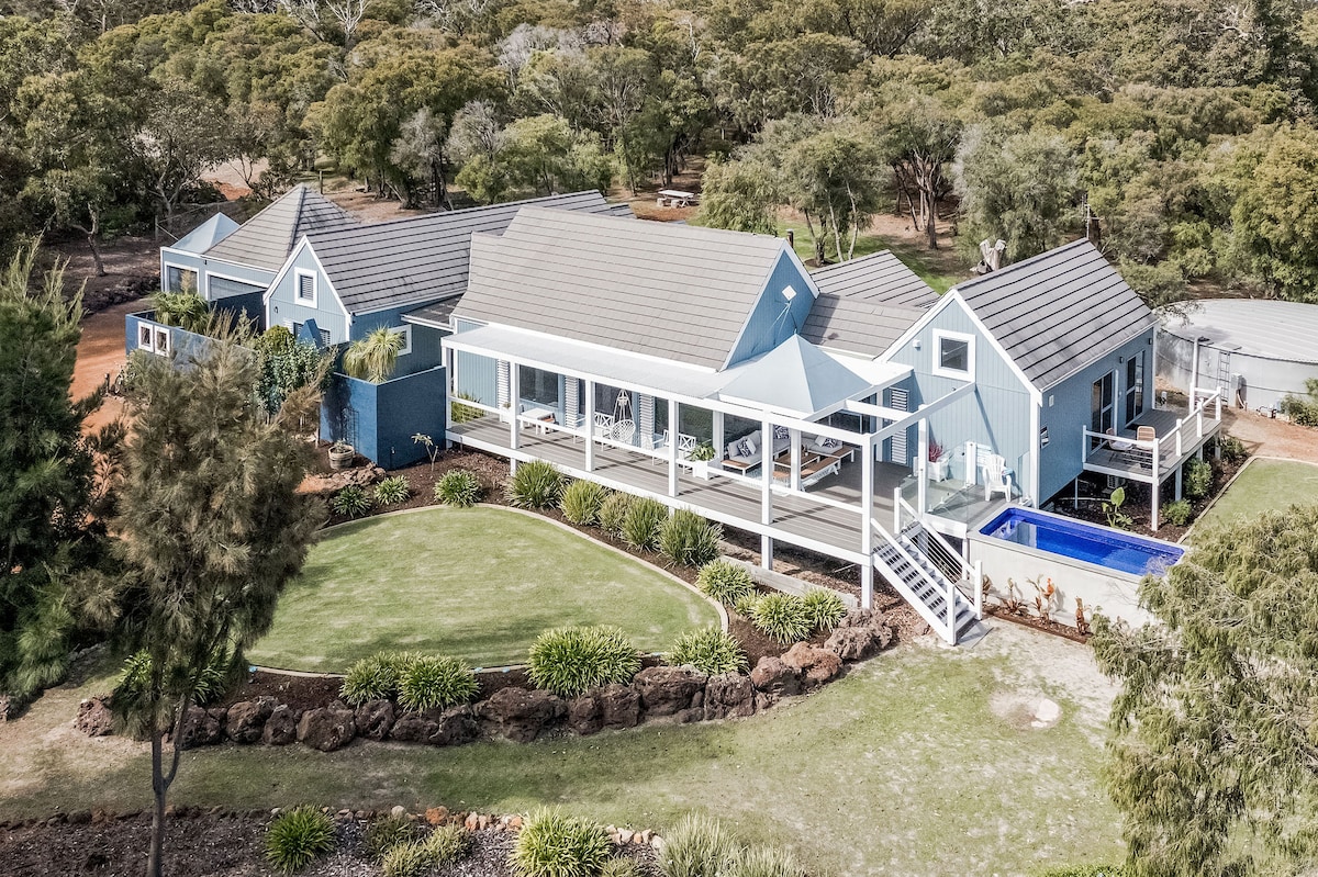 An aerial view captures a sprawling luxury estate featuring a large covered deck, surrounded by landscaped gardens. A plunge pool is visible to the right, with multiple buildings and an expansive lawn that blends harmoniously with the natural setting of trees and greenery.