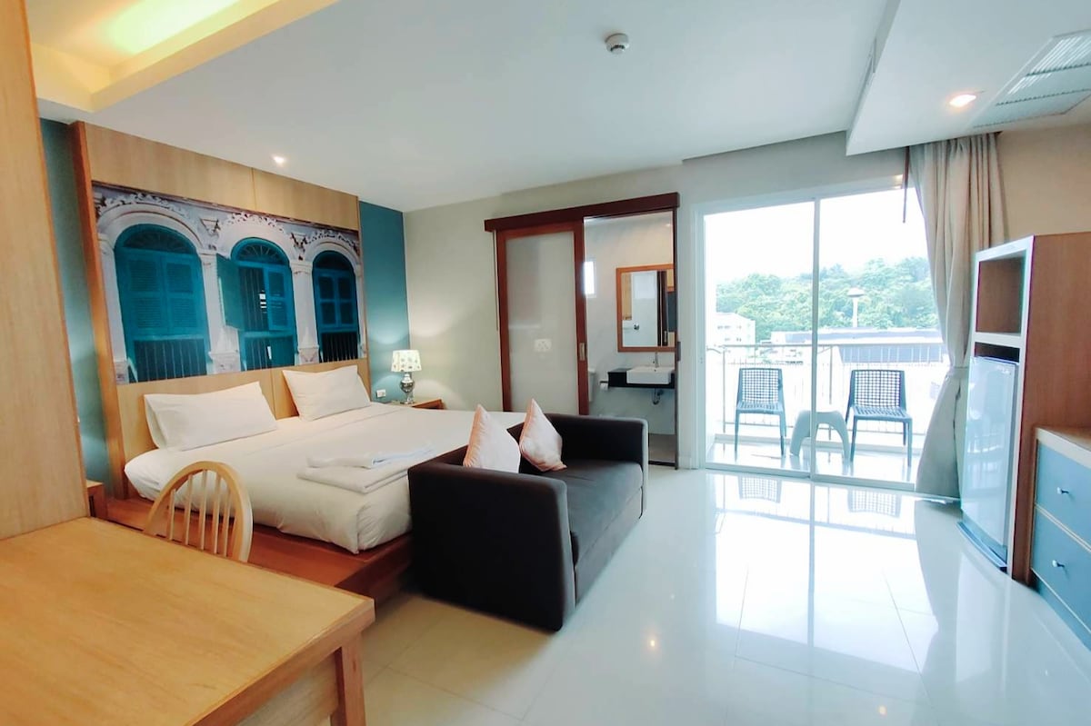 A modern guest room features a spacious bed adorned with neutral-toned linens. A comfortable sofa sits beside a wooden dining table. Large windows provide natural light, leading to a private balcony with outdoor seating. Decorative elements include blue accent walls and wall art.