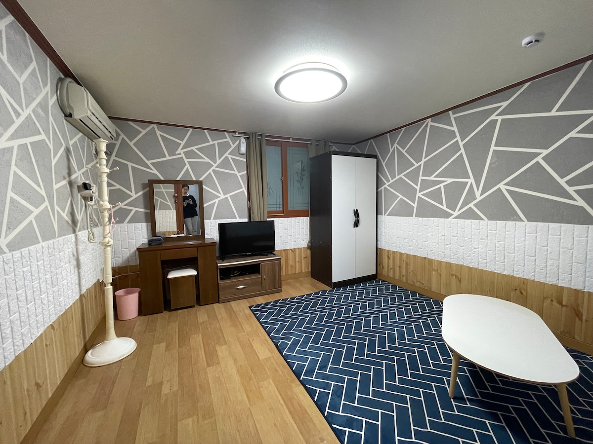 The room features modern geometric wall patterns alongside warm wooden accents. A low coffee table is centered on a blue patterned rug. A television unit and a white wardrobe are positioned against the walls. Natural light enters through the window, enhancing the neutral color scheme.