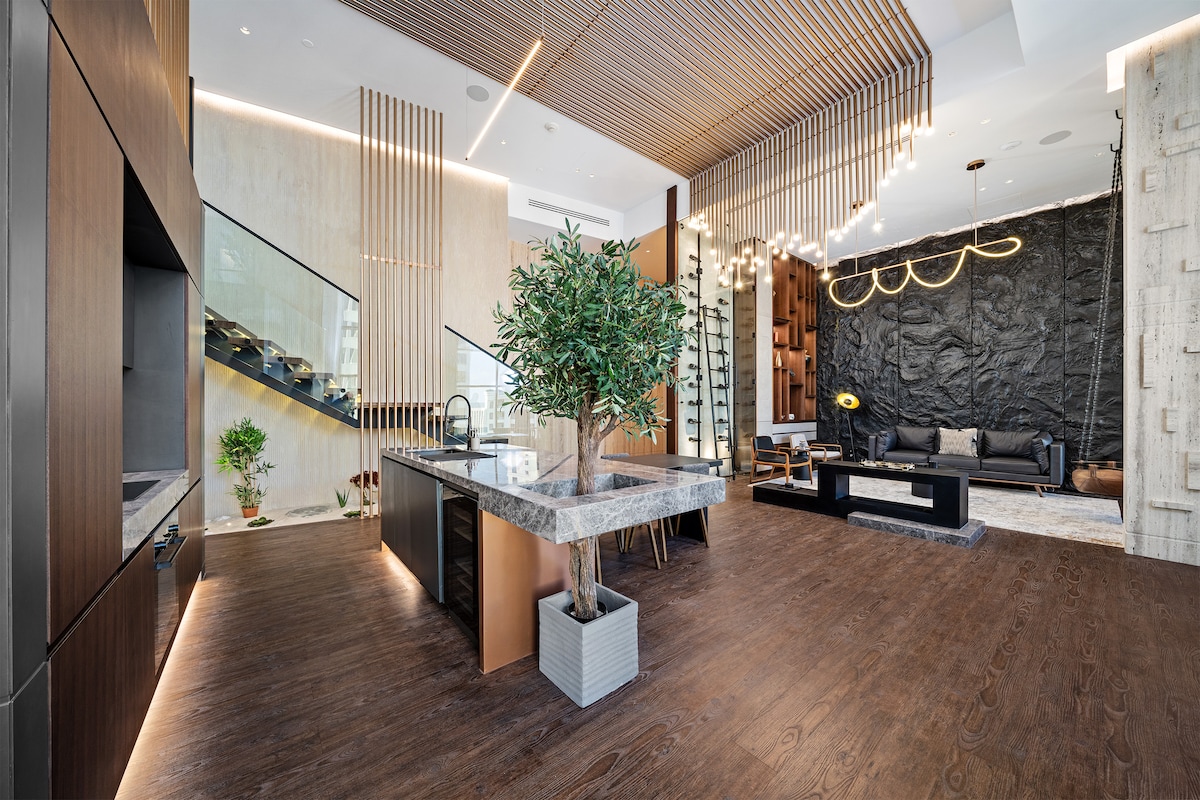 The spacious living area features modern architectural elements, including floor-to-ceiling windows and a stylish staircase. A central kitchen island with a marble countertop is complemented by elegant wooden cabinetry. An indoor plant adds a touch of greenery to the space, enhancing its contemporary design.