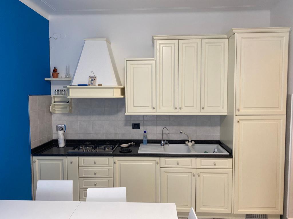 A kitchen area features cream-colored cabinetry with a black countertop. A gas stove and sink are visible, along with kitchen utensils. A blue accent wall complements the overall design. A white dining table and chairs are positioned nearby, creating a functional and welcoming space.