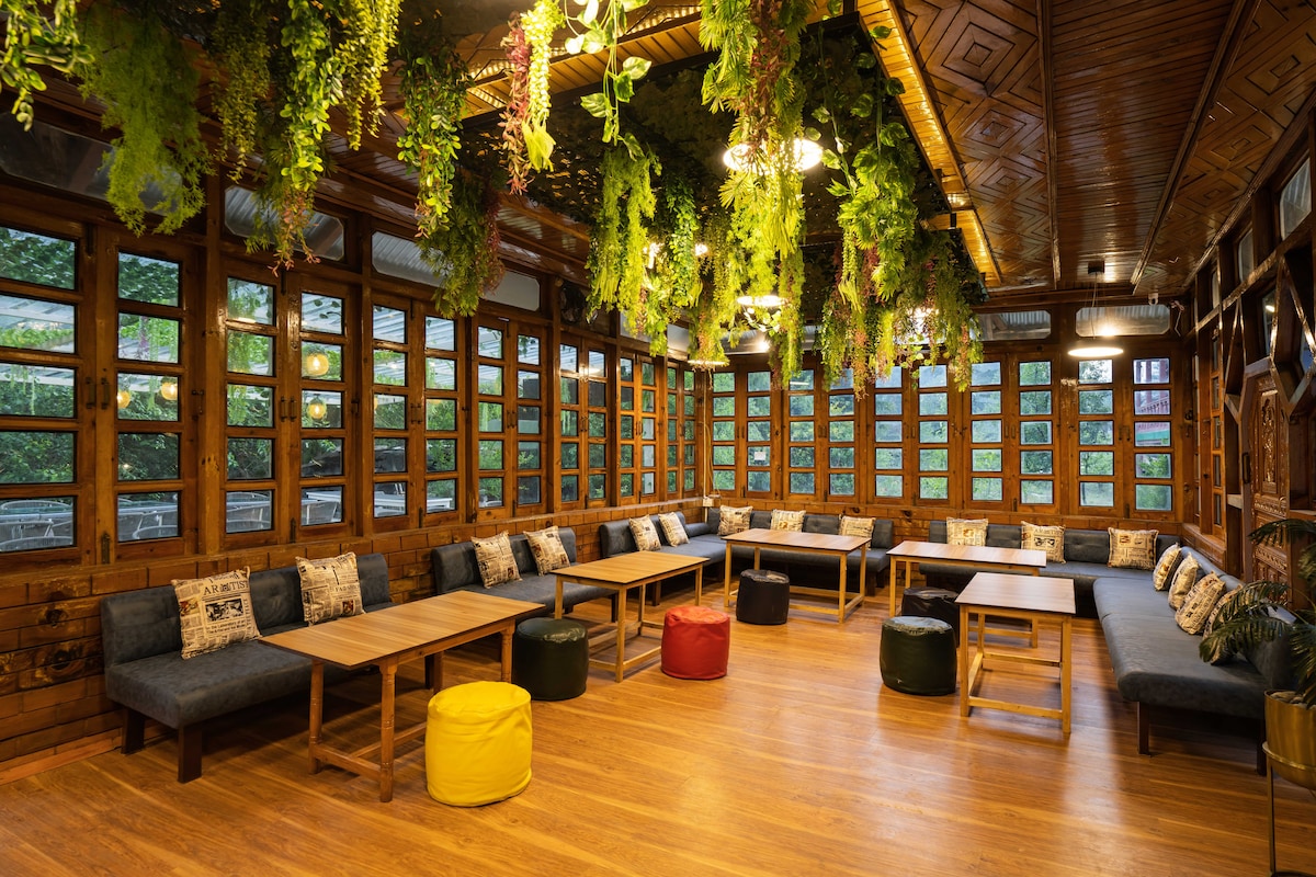 A spacious common room features a series of large windows allowing natural light to fill the space. Comfortable seating is arranged around several wooden tables, with colorful cushions and poufs adding a vibrant touch. Greenery hangs from the ceiling, enhancing the lively atmosphere.