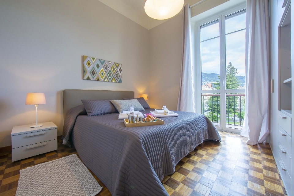 A spacious bedroom features a large bed adorned with a neutral-colored blanket. A bedside table with a lamp sits beside the bed. Natural light enters through a large window, offering views of greenery outside. A colorful piece of wall art adds a touch of character.