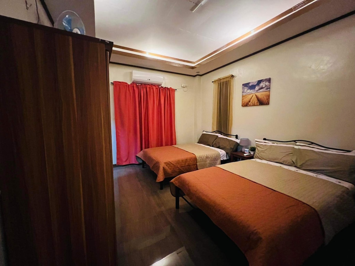 A cozy sleeping area is presented with two spacious beds covered in soft blankets. A wardrobe stands against the wall, and curtains in warm tones frame the window. An air conditioning unit provides comfort, while a piece of wall art adds a subtle decorative touch.