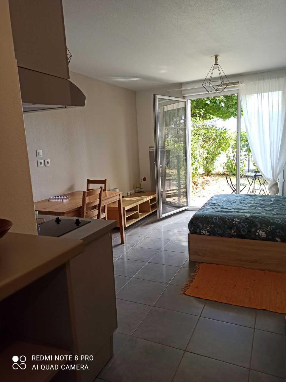 A well-lit studio features large glass doors that open to a small private garden. A cozy seating area is visible, alongside a kitchenette with a stove. The space is decorated with soft colors and a patterned bedspread on the spacious bed, creating a relaxing atmosphere.