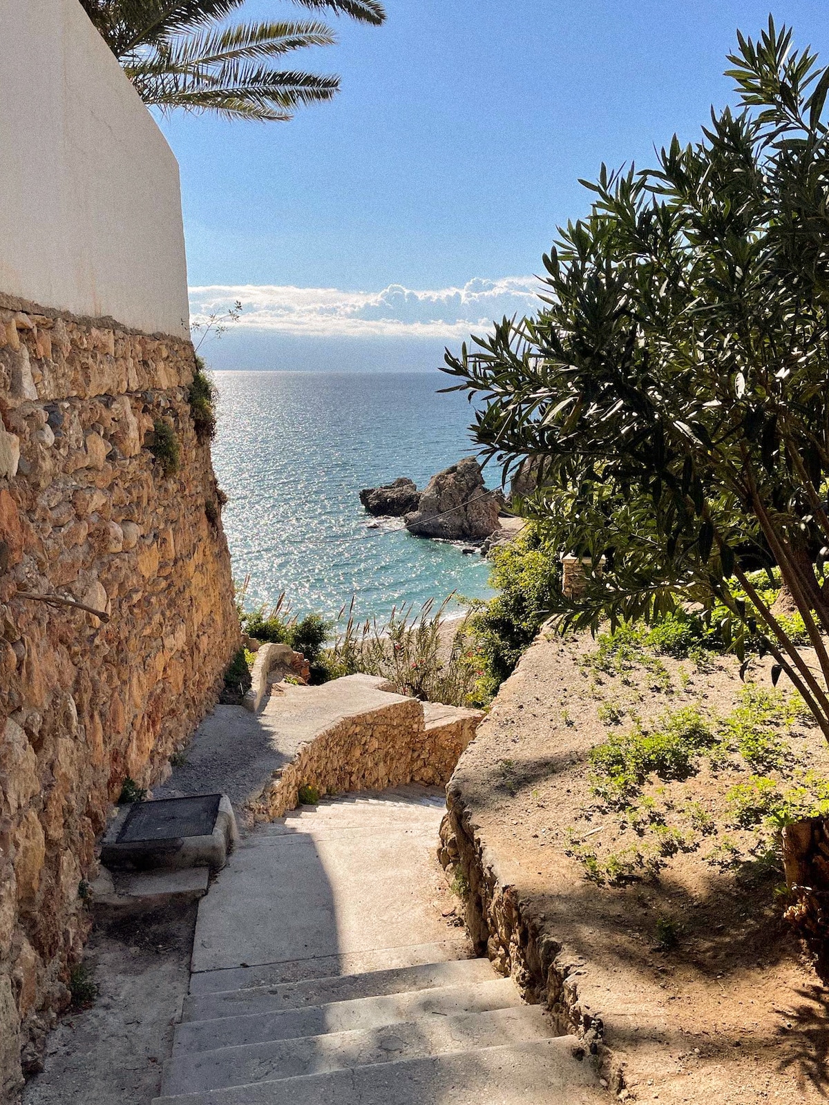 A stone pathway descends towards the sea, framed by lush greenery on both sides. The sunlight reflects off the water, creating a serene scene. In the distance, a rocky outcrop is visible along the shoreline, enhancing the natural beauty of the surroundings.