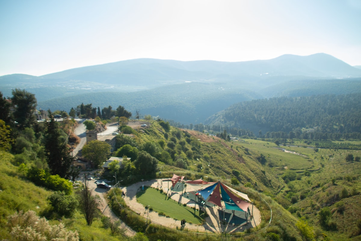 A scenic landscape unfolds, featuring rolling hills and lush greenery under a clear blue sky. A colorful playground with a unique canopy design can be seen at the base of the hills, offering a recreational space surrounded by nature.