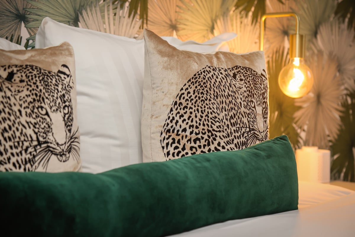 The bed is adorned with decorative cushions featuring leopard designs and a plush green bolster. A warm light emanates from a stylish bedside lamp, illuminating the surrounding leafy wallpaper that creates a serene ambiance.