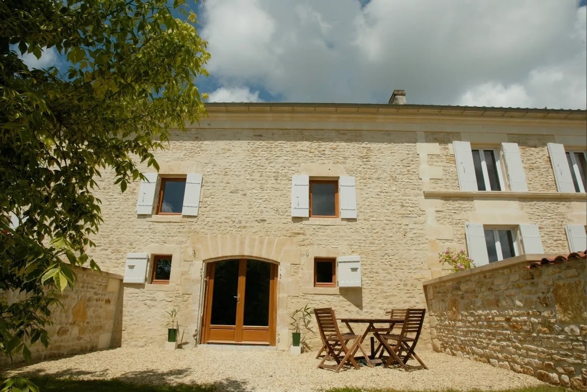 The exterior of the gîte showcases a charming stone facade with wooden shutters. A gravel area is furnished with a small table and folding chairs, surrounded by greenery. Large windows provide natural light to the interior, enhancing the inviting appearance.
