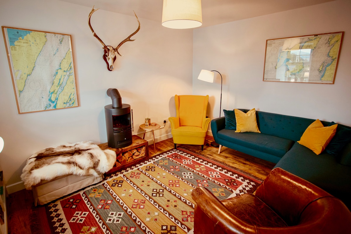 The cozy living area features a vibrant blue sofa and a mustard yellow armchair, accented by decorative throw pillows. A decorative rug covers the floor, while a wall-mounted deer antler adds a rustic touch. Natural light illuminates the space from a nearby window.