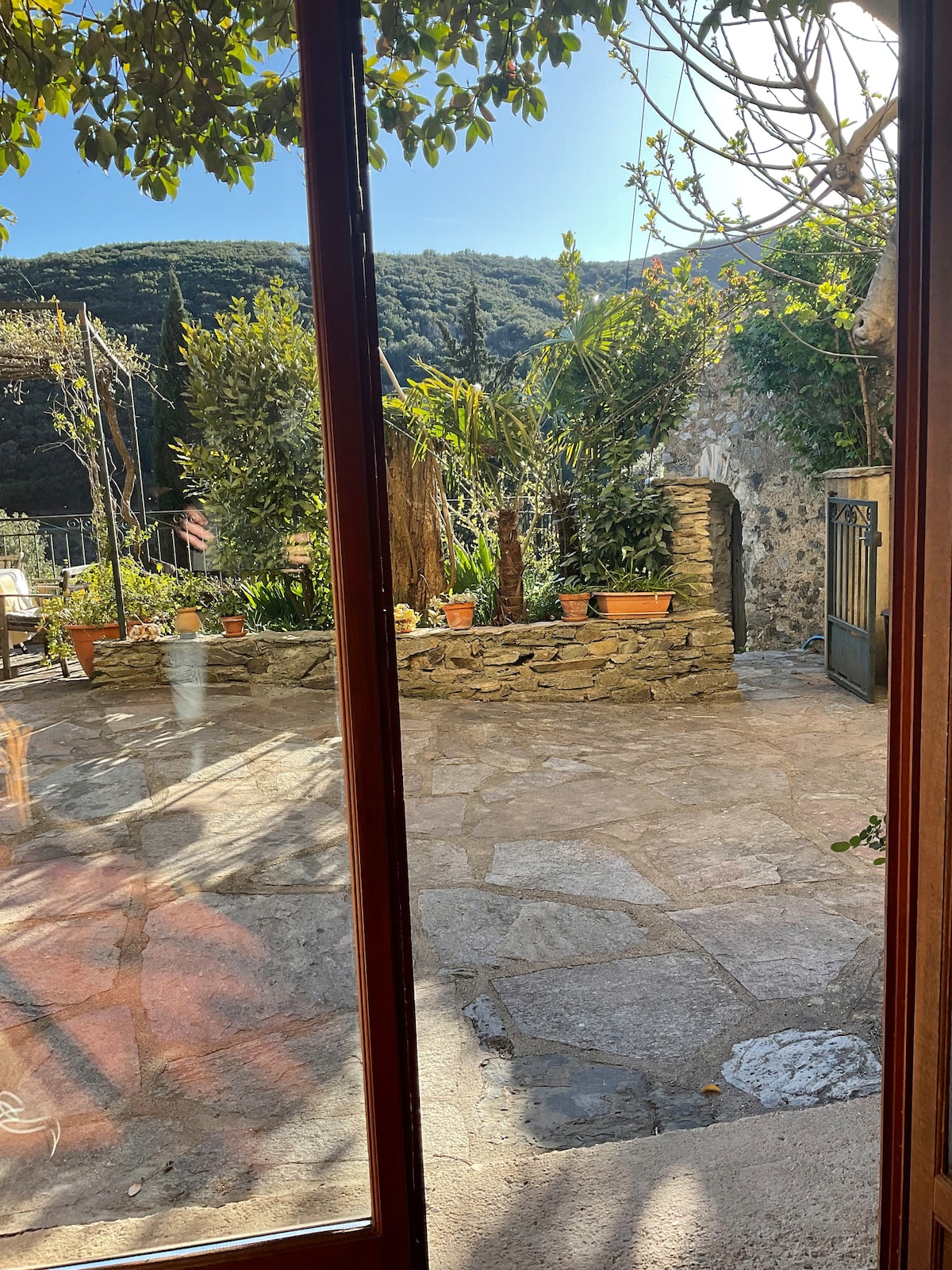 A stone patio is visible through a large wooden window frame, featuring various potted plants arranged throughout the outdoor space. In the background, lush, tree-covered mountains can be seen, providing a serene natural setting.