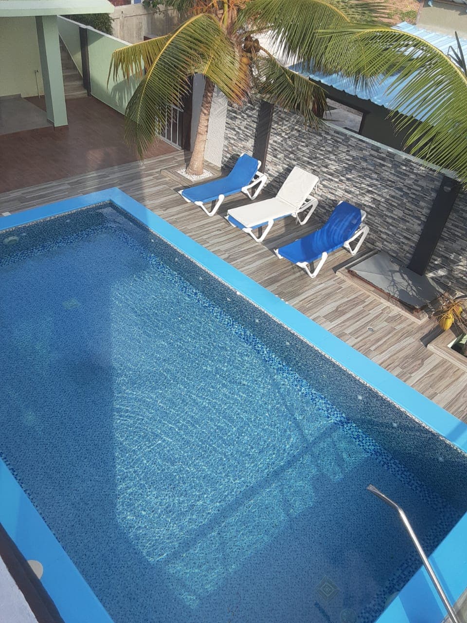 A backyard pool area features a rectangular pool surrounded by light-colored decking. Two white lounge chairs with blue cushions are positioned nearby. Lush greenery and a stone accent wall provide a natural backdrop, while sunlight reflects off the water's surface.