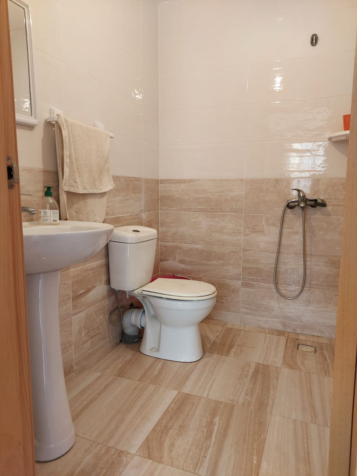 A clean and well-maintained bathroom is shown, featuring light-colored tiles and simple fixtures. A white toilet and a pedestal sink are positioned alongside a glass shower with a handheld showerhead. Towels are hung neatly, and a small shelf holds toiletries.
