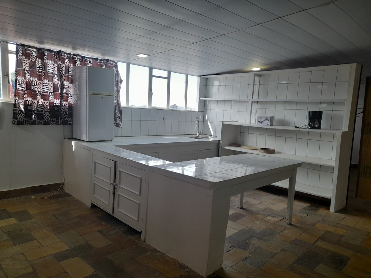 The kitchen features a spacious countertop surrounded by white cabinetry. Natural light enters through large windows adorned with patterned curtains. A refrigerator and coffee maker are visible, complemented by a tiled floor that adds texture to the space.
