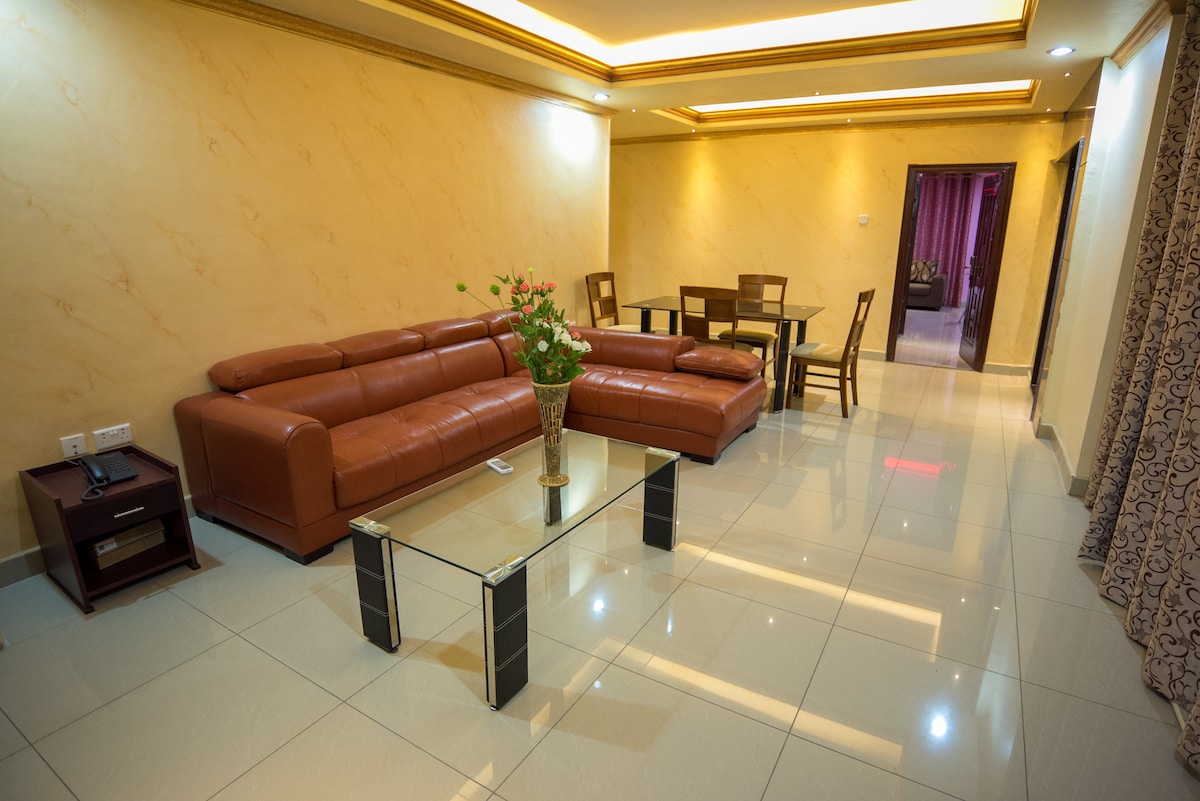 A spacious living area features modern, brown leather seating and a glass coffee table at the center. A dining table with wooden chairs is visible, and soft ambient lighting highlights the room's neutral tones. Decorative curtains frame the windows, adding a touch of elegance.