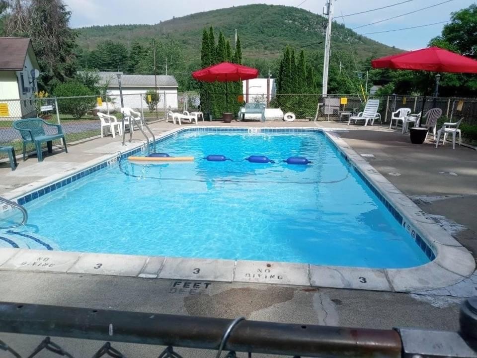 A clear swimming pool is surrounded by a fenced area, featuring several lounge chairs and red umbrellas providing shade. The backdrop includes a gently sloping green hill, contributing to a serene outdoor setting.