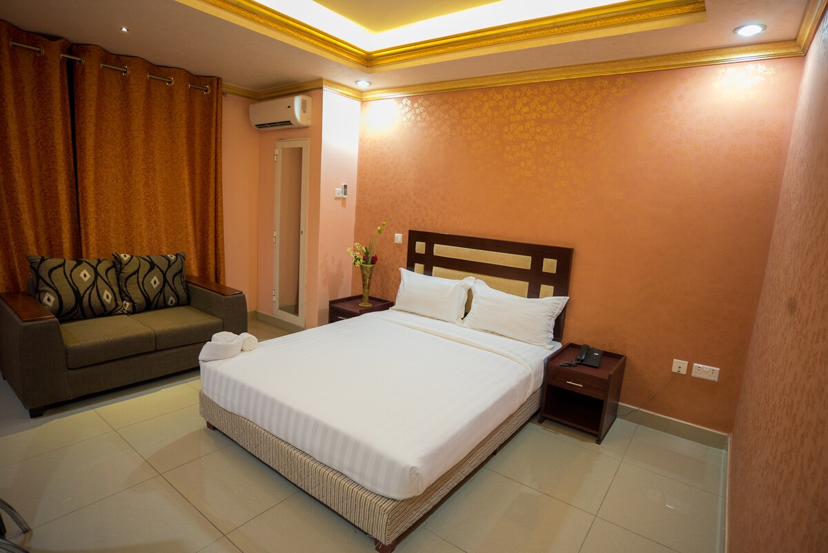 The room features a spacious bed with crisp white linens against a subtle pink wall. A cozy sofa sits nearby, complemented by elegant curtains. A bedside table and wall-mounted phone are visible, along with soft lighting that enhances the overall space.
