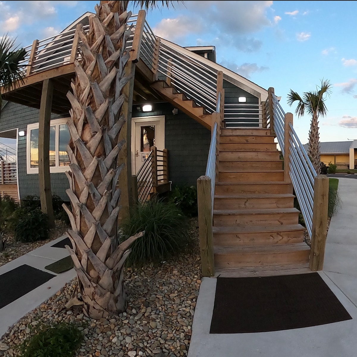 An inviting entrance is showcased, featuring a wide wooden staircase with a sturdy railing. Lush greenery and palm trees frame the pathway, which is bordered by decorative stones. The exterior of the building is finished in earthy tones, with soft lighting illuminating the steps.
