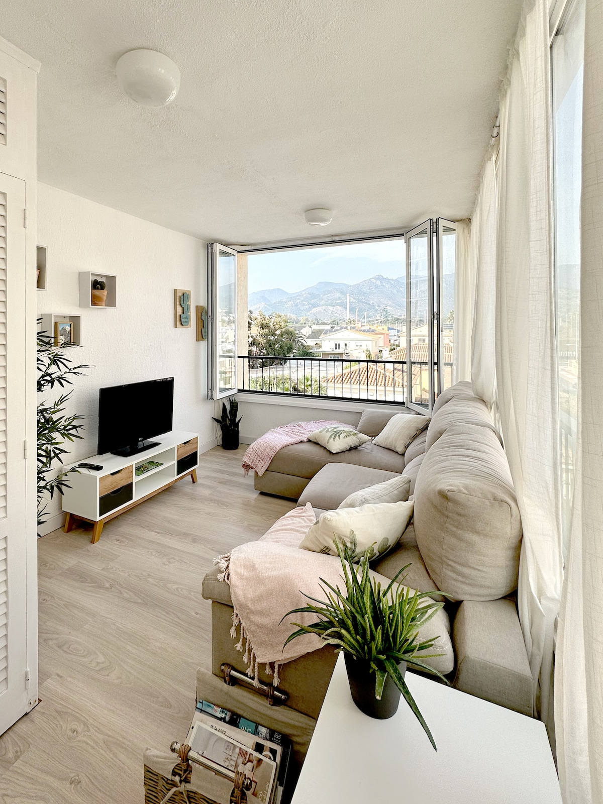 A bright living area features large windows that open to stunning mountain views. A comfortable sectional sofa is complemented by soft cushions. A small green plant adds a touch of greenery, while a television is mounted against the wall, enhancing the spacious ambiance.