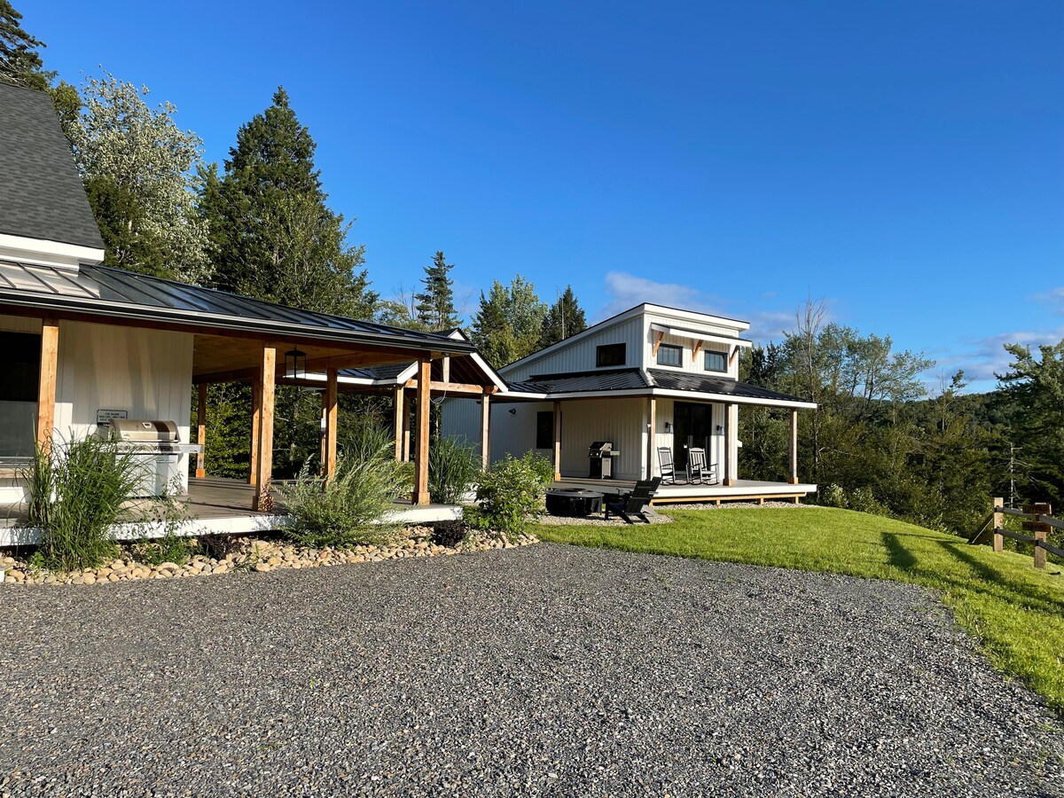 A well-maintained outdoor area showcases a gravel driveway leading to the Guest Cottage. The modern cottage displays a two-story design with a large porch, while lush greenery surrounds the property. The serene landscape extends in the background, offering a glimpse of the natural surroundings.
