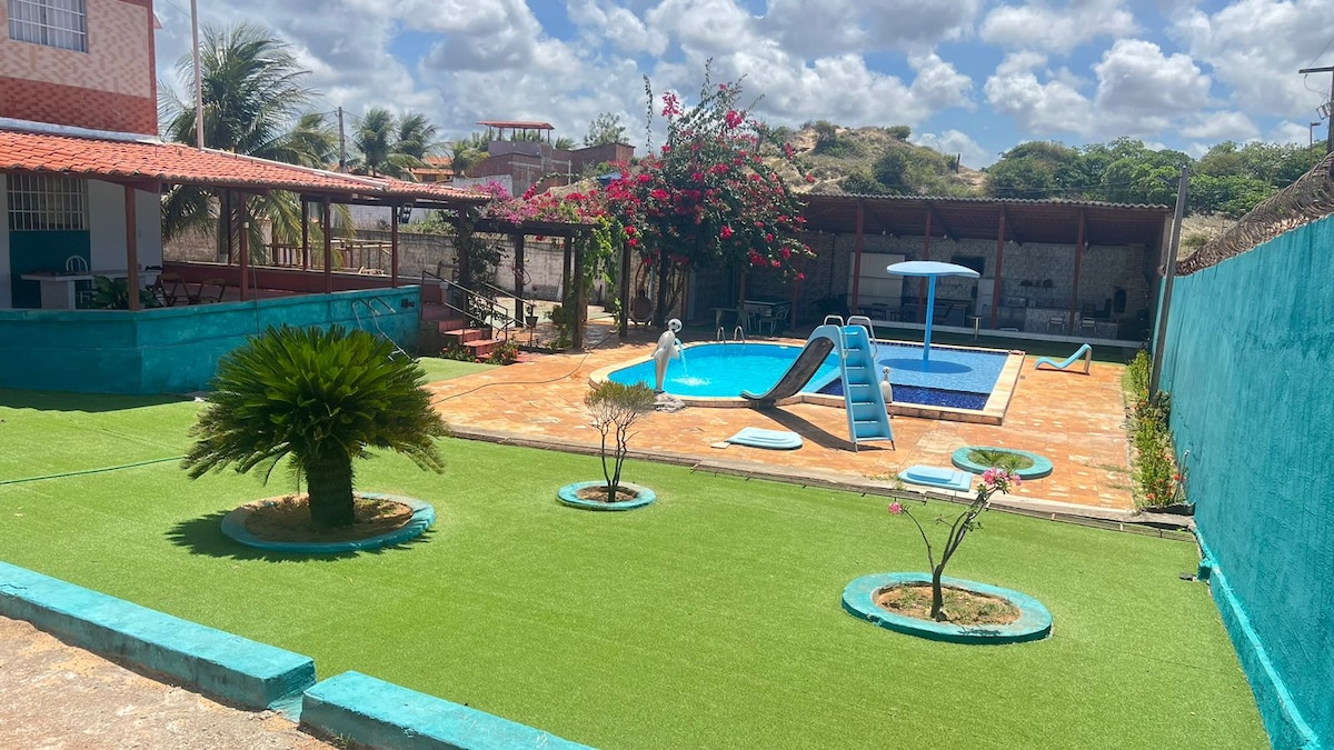 The outdoor area features a well-kept pool surrounded by vibrant green grass, complemented by landscaped flower beds. A separate shallow pool for children is included, alongside a pool slide. A shaded seating area and colorful foliage enhance the tranquil setting.