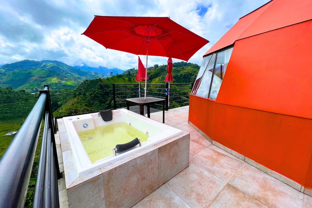 An outdoor terrace features a jacuzzi surrounded by a landscaped view of mountains. Two red umbrellas provide shade over a black table and chairs. A vibrant red structure sits nearby, complementing the natural scenery.