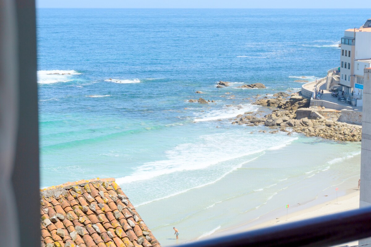 A view of the coastline is presented, featuring gentle waves lapping at the sandy shore. The scene includes rocky formations and a clear blue sea. Nearby buildings are visible, positioned along the waterfront, enhancing the tranquil seaside environment.