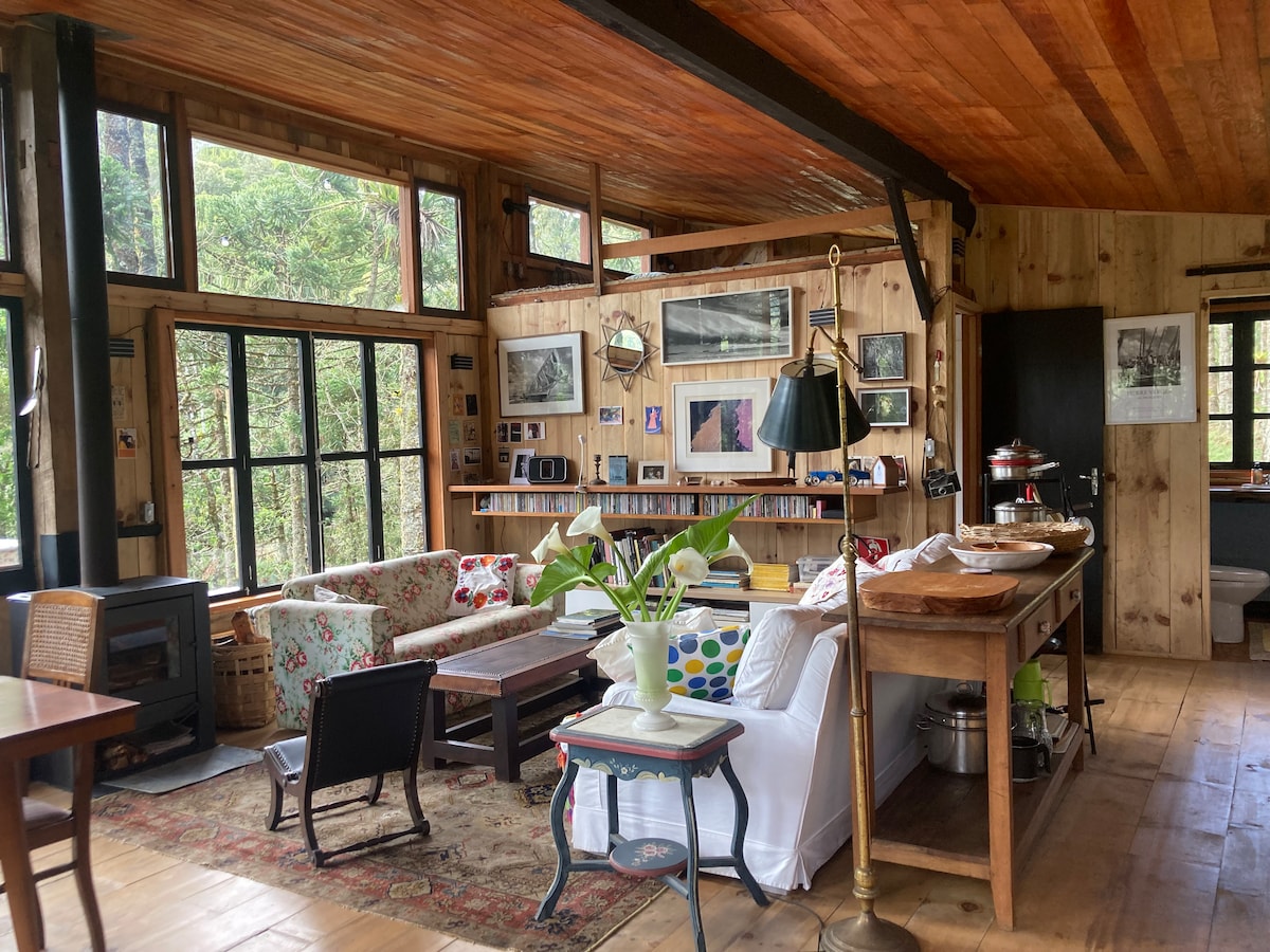 The living area showcases a mix of cozy furnishings, including a floral-patterned sofa and a white slipcovered loveseat. Large windows allow natural light to fill the space, providing views of the surrounding nature. A wooden dining table, lamps, and decorative art enhance the inviting ambiance.