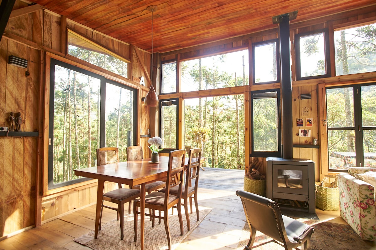 A warm and inviting dining area features a large wooden table surrounded by six chairs, with large windows showcasing views of the surrounding forest. A wood stove stands in the corner, providing a cozy focal point within the space.