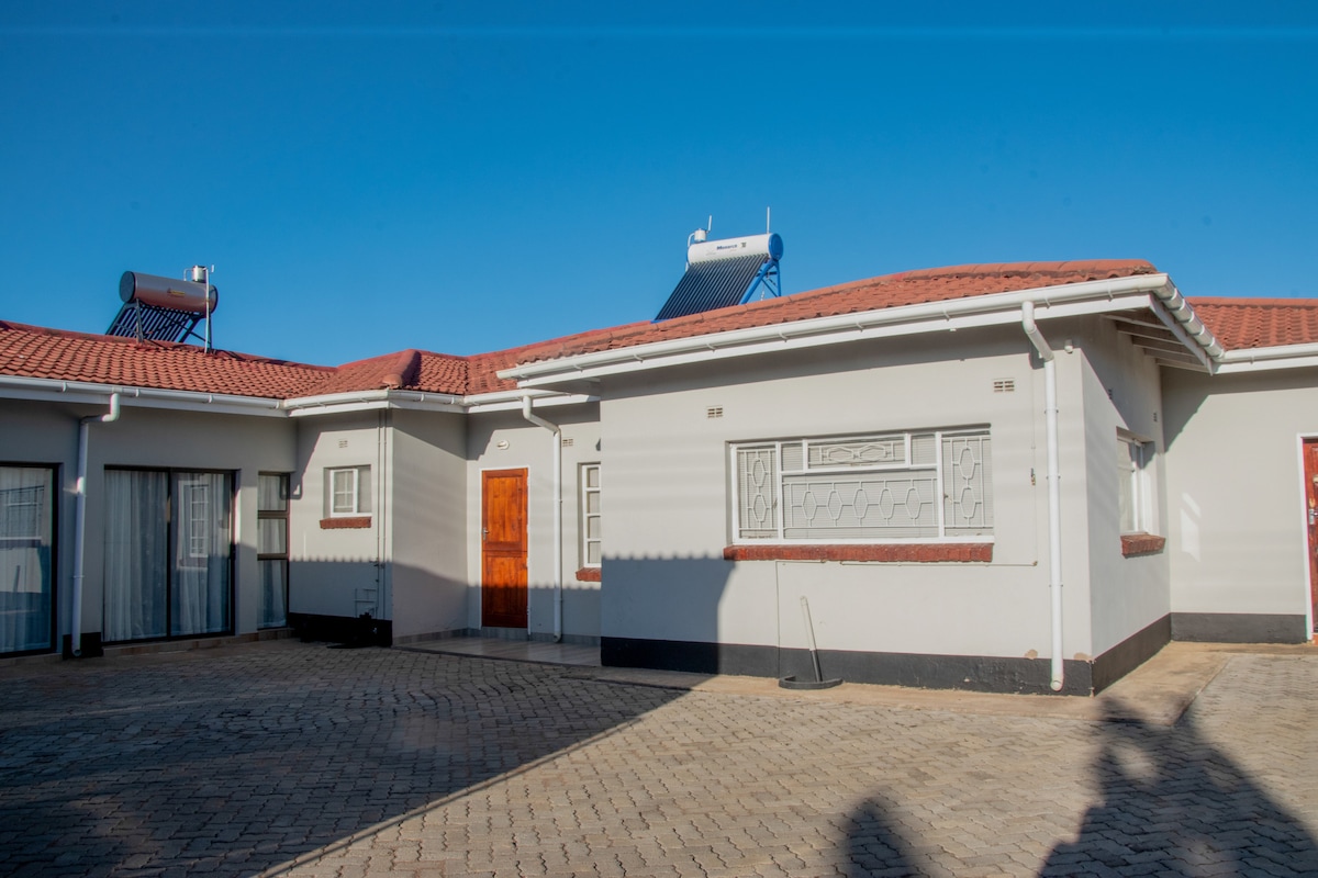 A single-story building showcases a flat roof with solar panels. The exterior is finished in light grey with contrasting dark trim. A wooden door is featured prominently, alongside large glass doors that allow ample natural light into the interior.