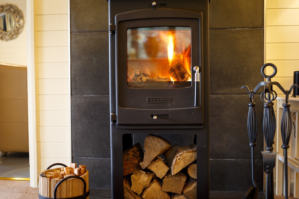 A wood-burning stove is featured, with flames visible behind the glass door. Stacked logs are neatly arranged beneath the stove. A basket holding firewood is positioned nearby, contributing to a cozy atmosphere.