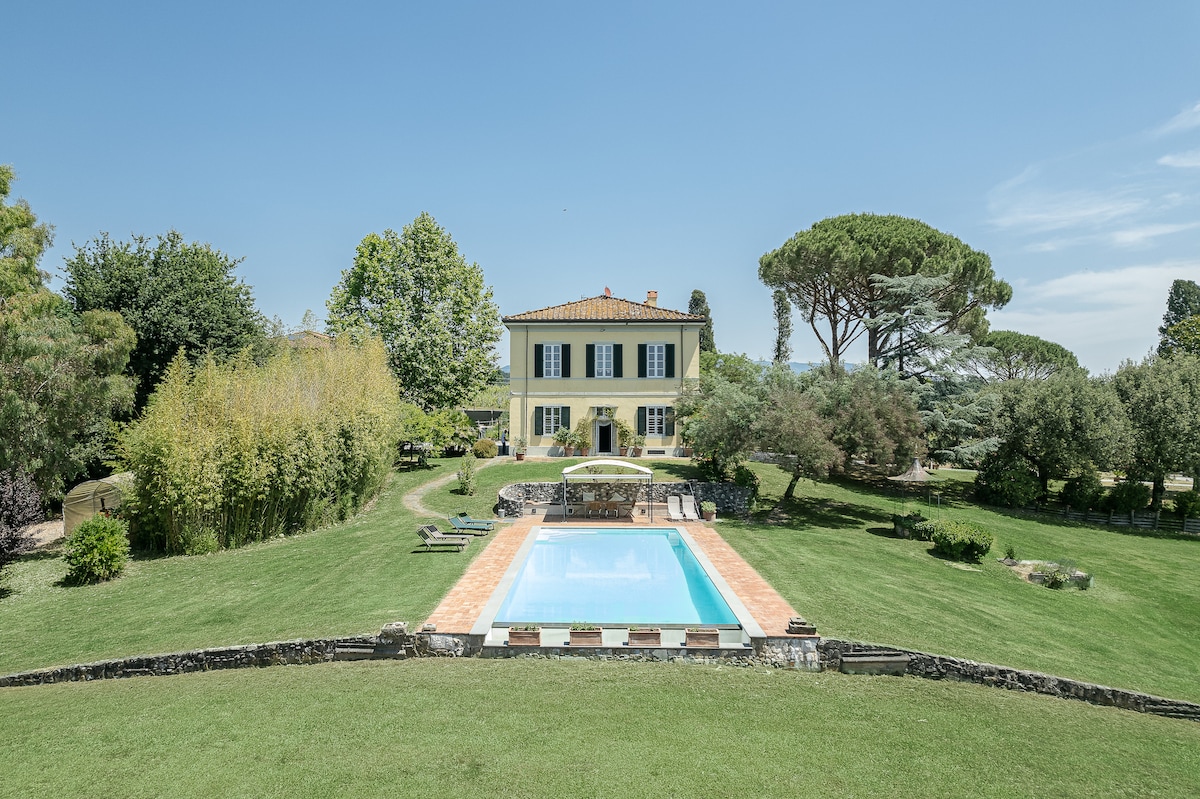 The historic villa is framed by lush greenery, with a tranquil swimming pool situated at the forefront. The pool's clear water reflects the surrounding trees and manicured lawn, while the villa's elegant façade features a broad terrace and large windows overlooking the expansive garden.
