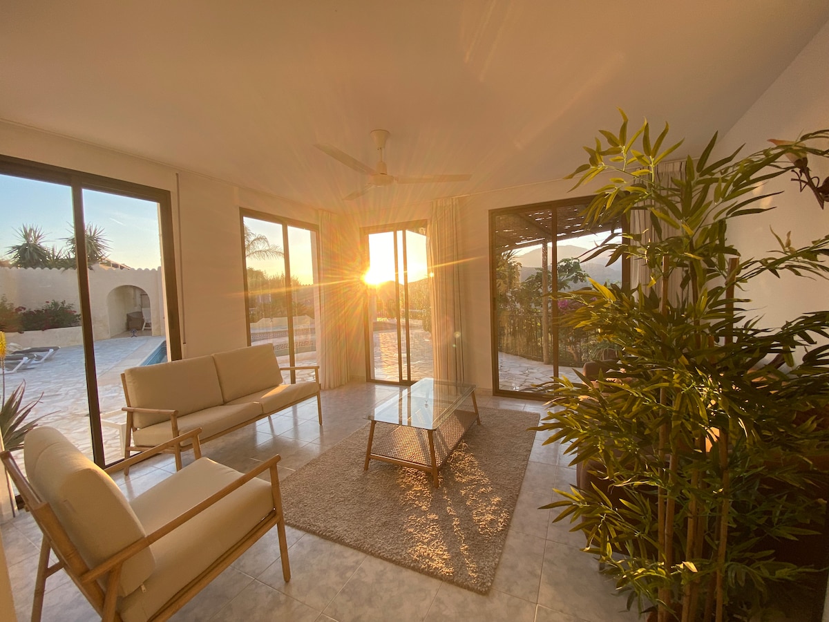 A sunlit living area features large sliding glass doors that open to a patio, allowing natural light to fill the space. Minimalist furniture includes light-colored armchairs and a glass coffee table, with a soft rug underfoot. A green plant adds a touch of nature.