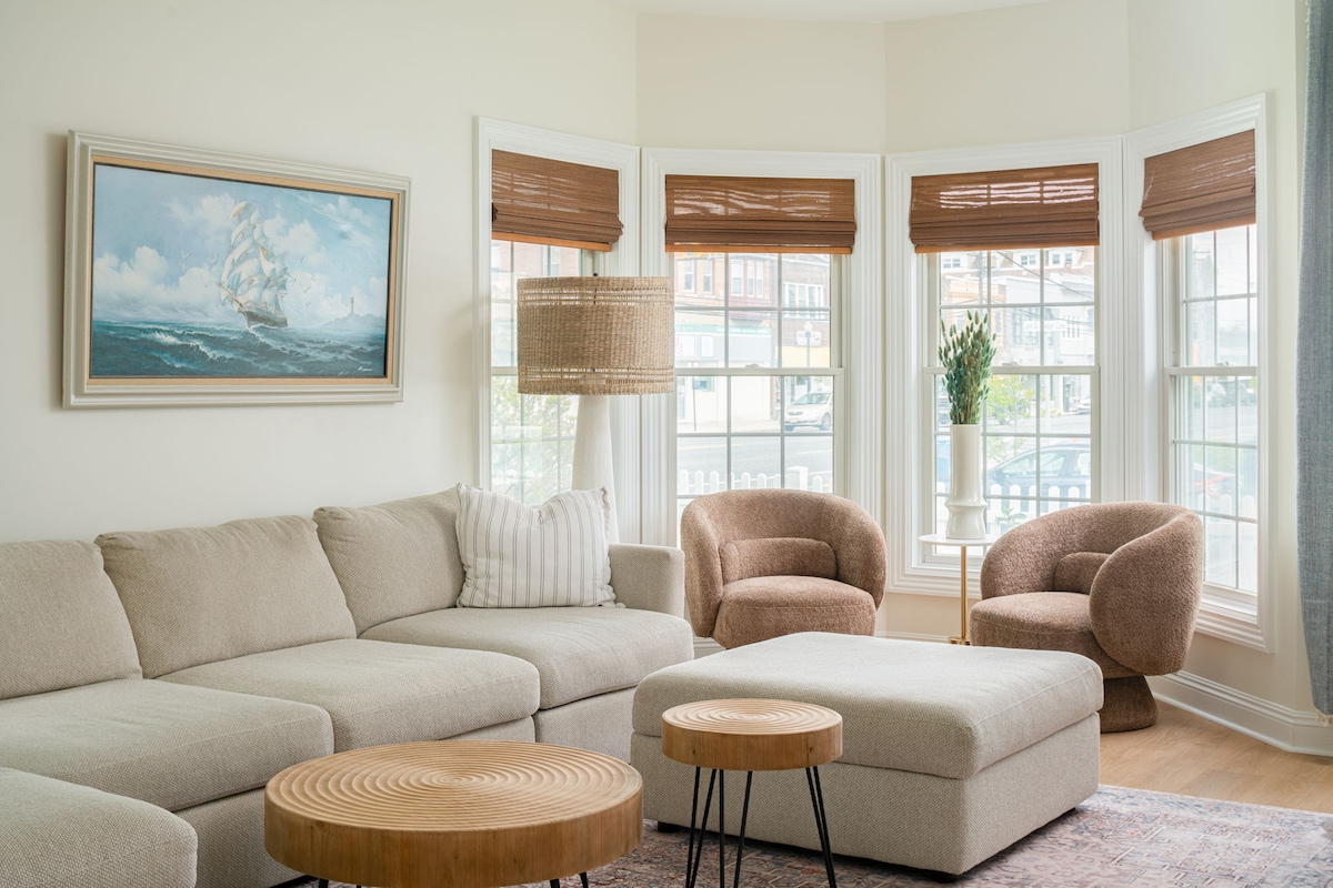 A light-filled living room is highlighted by a soft, neutral sectional couch and two accent chairs. A round coffee table sits between them, and art featuring a sailing ship adorns the wall. Large windows with wooden shades offer views of the outside.