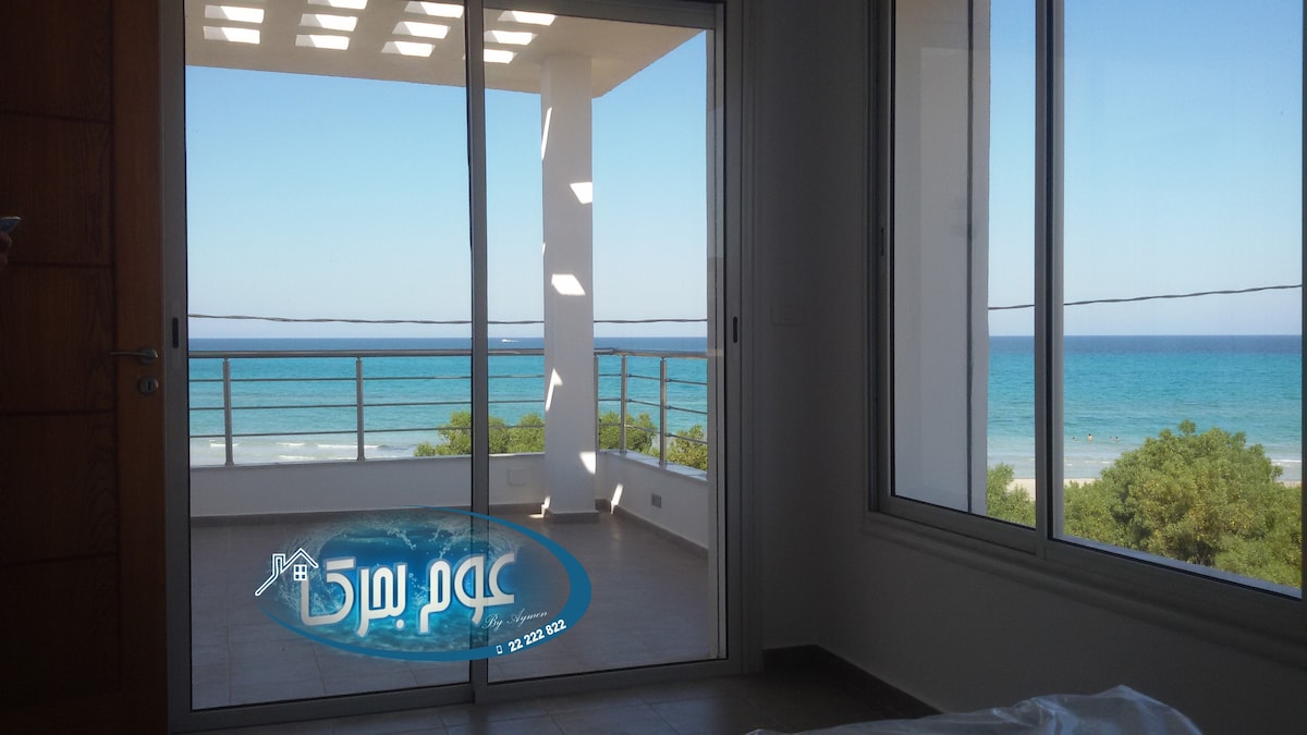 A spacious room features large sliding glass doors, presenting unobstructed views of the sea. The open layout allows natural light to flow in, illuminating the minimal decor. The shoreline and greenery can be seen through the glass, enhancing the tranquil environment.