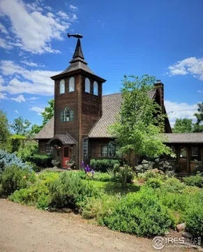 A charming farmhouse is surrounded by a variety of lush greenery and colorful flowers. The structure features a distinctive turret and a sloped roof, complemented by wooden siding. The clear blue sky highlights the inviting exterior and beautifully landscaped grounds.