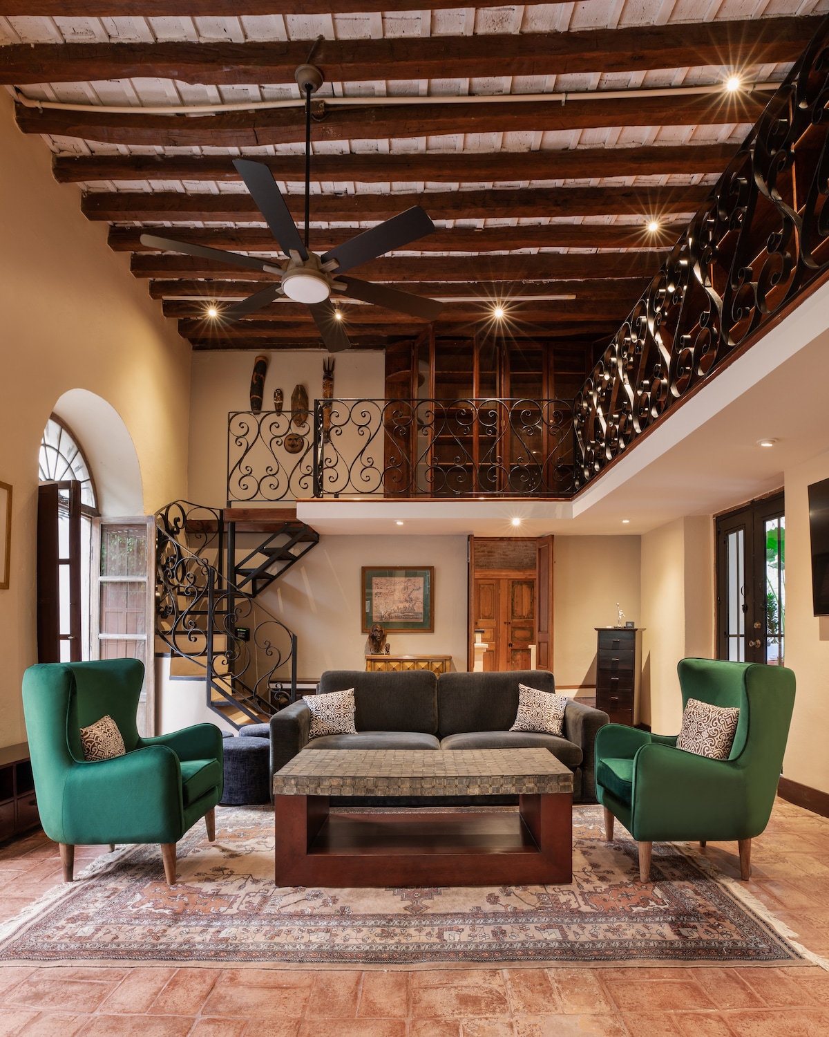 A spacious living area features high wooden ceilings, an ornate iron railing, and natural light streaming through large windows. Two green armchairs frame a stylish coffee table, while a plush sofa invites relaxation. A decorative area rug enhances the room's warm and welcoming atmosphere.