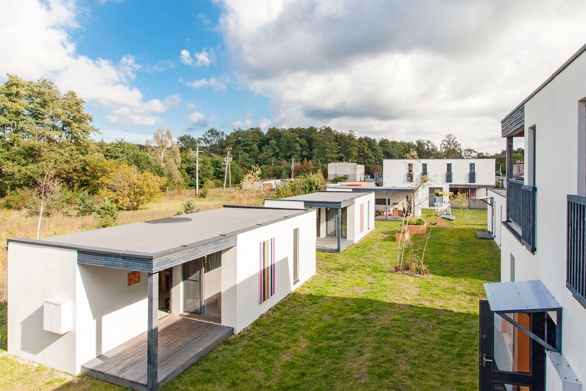 A collection of individual cottages is arranged around a spacious green lawn, surrounded by trees and nature. The architecture features flat roofs and minimalist design, with colorful accents on some walls. A play area can be seen in the distance, enhancing the family-friendly environment.