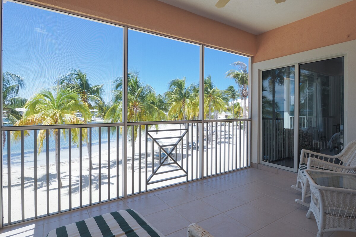 A screened-in balcony features white wicker seating overlooking swaying coconut palms and a sandy beach. Natural light floods the space, illuminating the tile floor. Sliding glass doors provide easy access to the coastal view, enhancing the serene environment.